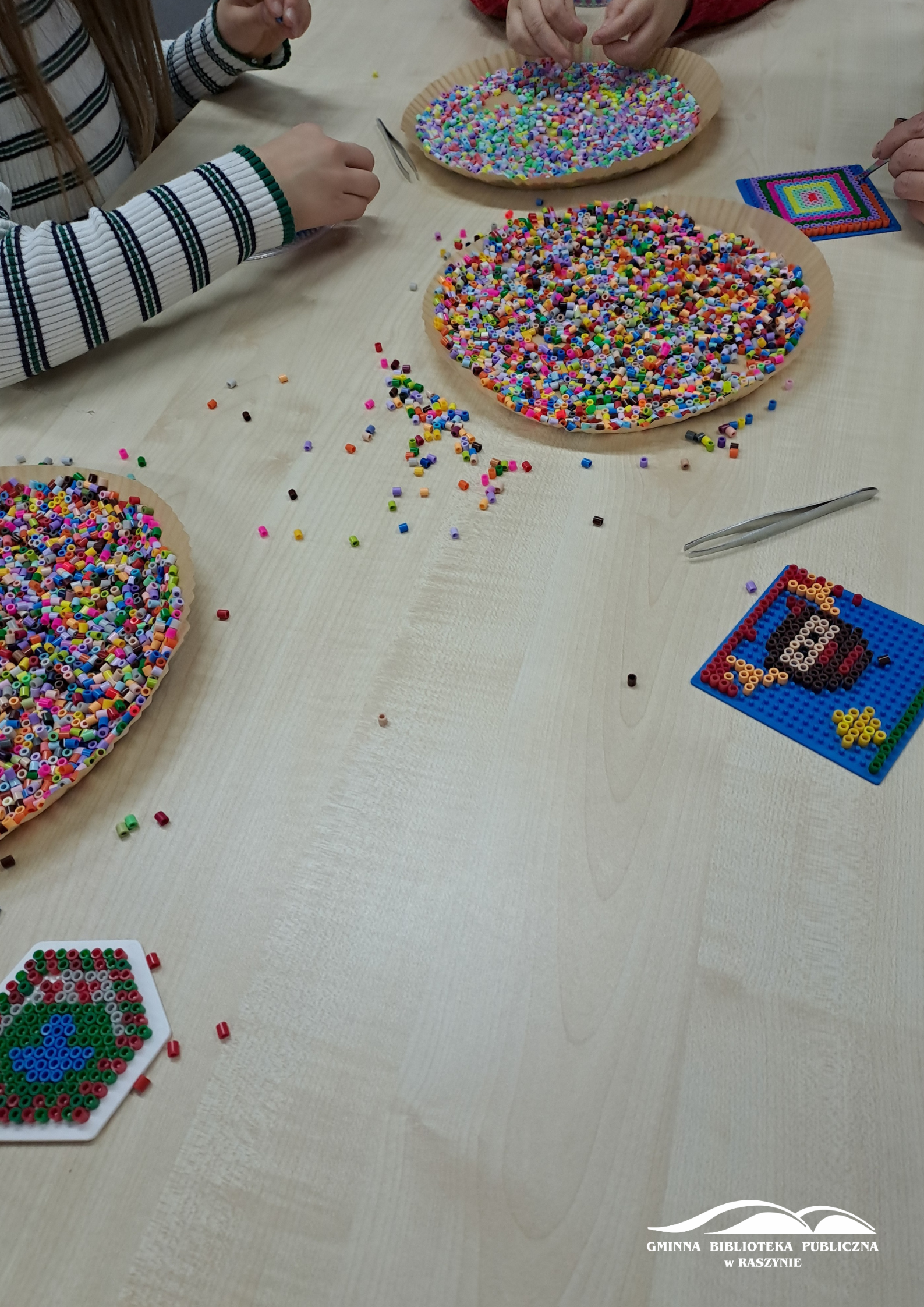 Zbliżenie na stół - widać powstające prace i dwie miseczki z koralikami