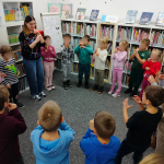 Dzieci wraz z bibliotekarką ustawione w koło śpiewają misiową piosenkę