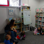 Dzieci siedzą na dywanie w dziale dla najmłodszych, bibliotekarka pokazuje na planszę z wizerunkami misiów z bajek