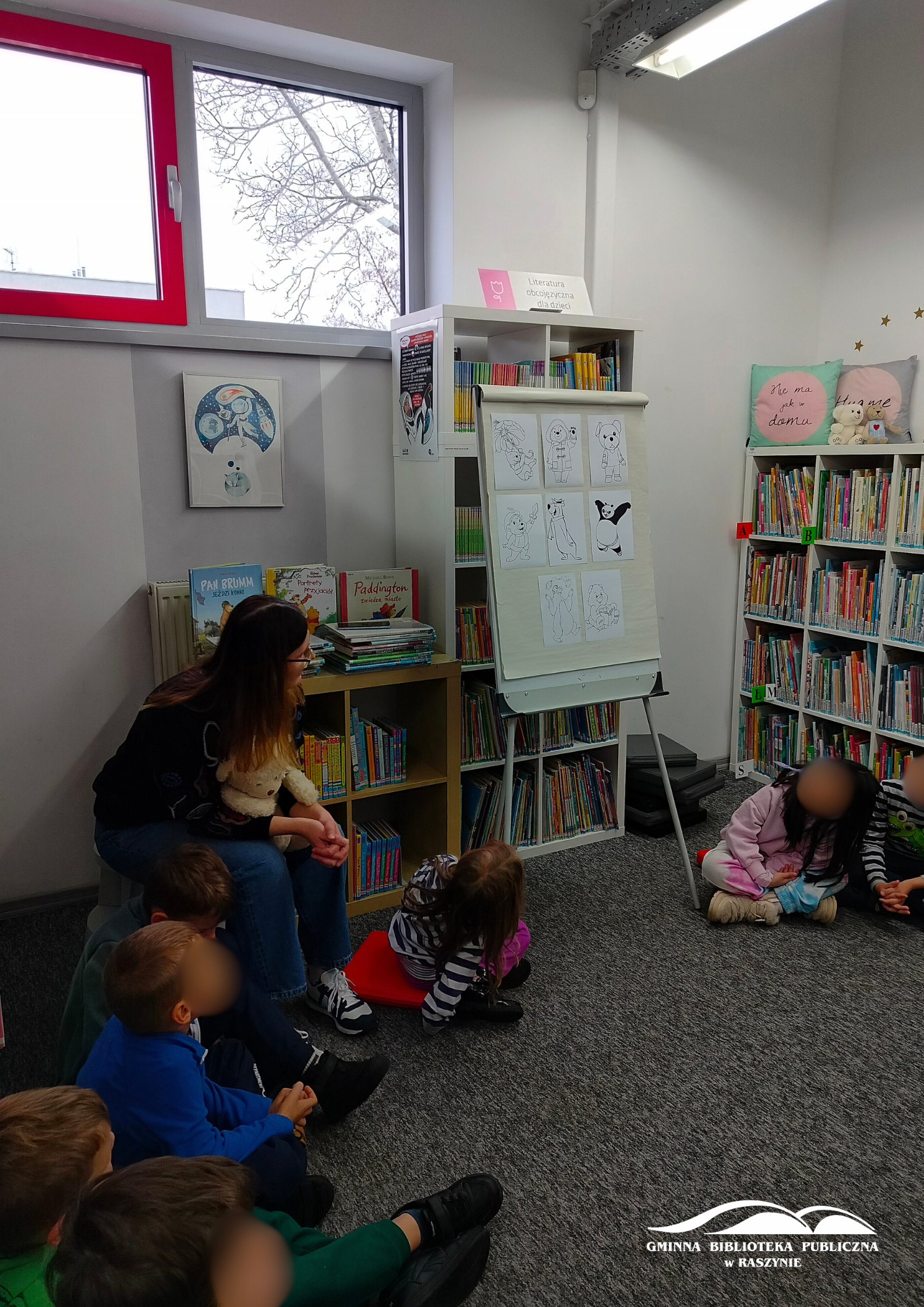 Dzieci siedzą na dywanie w dziale dla najmłodszych, bibliotekarka pokazuje na planszę z wizerunkami misiów z bajek