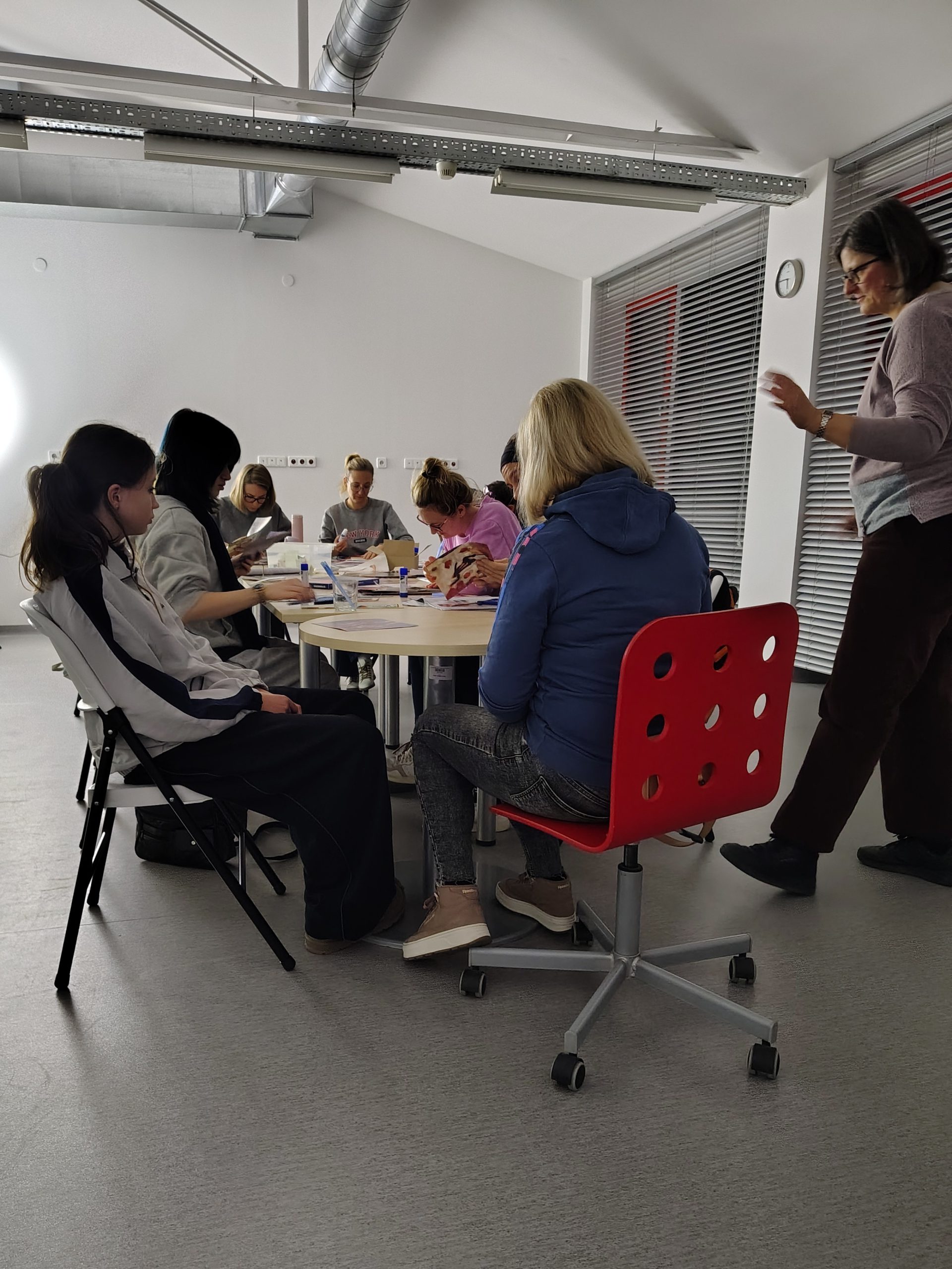 Uczestniczki warsztatów kolażowych siedzą przy stole, obok stoi prowadząca, widać rozłożone papiery i gazety