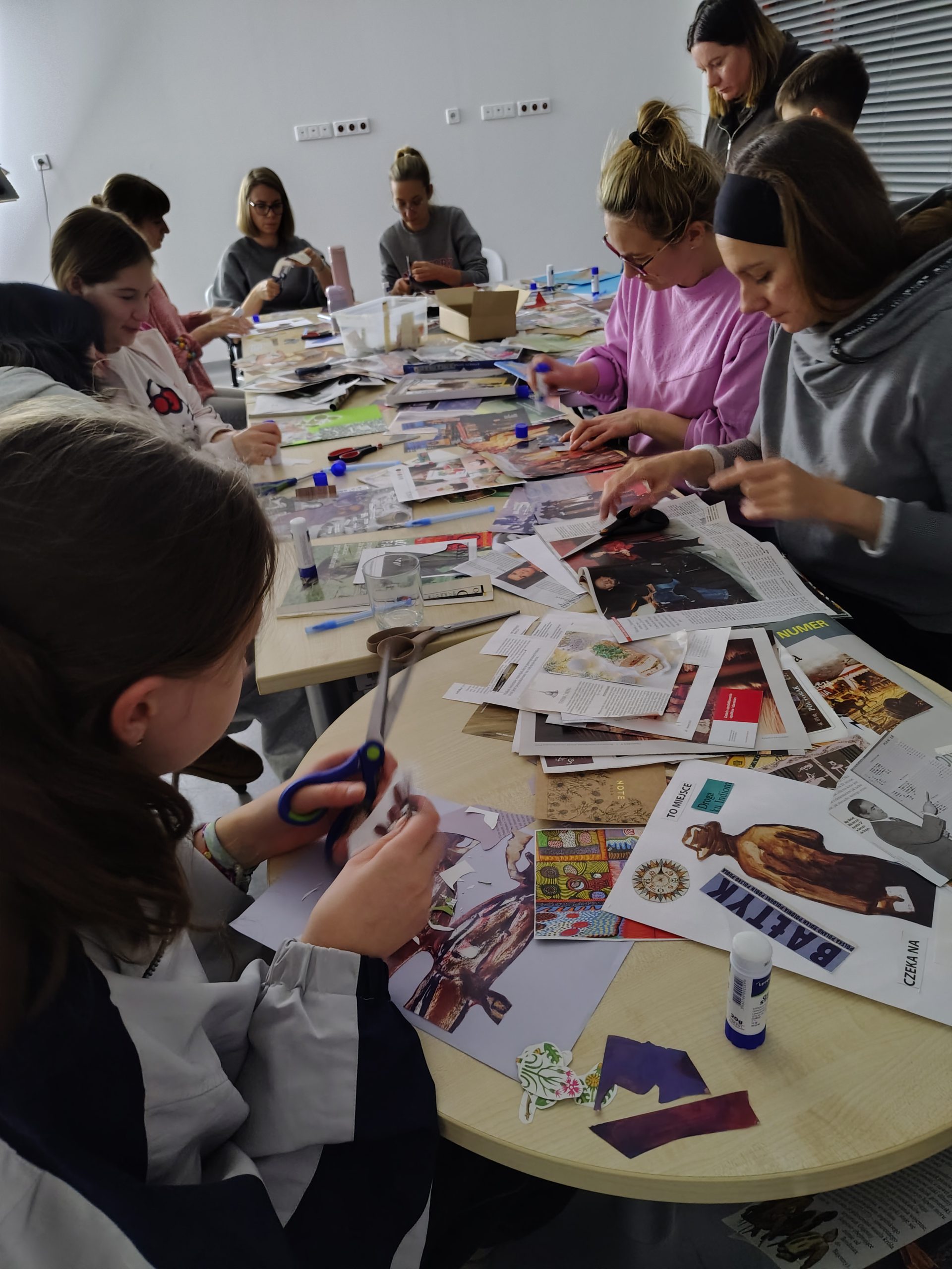Uczestniczki warsztatów kolażowych podczas pracy - zbliżenie na stół z materiałami, widać ręce z nożyczkami