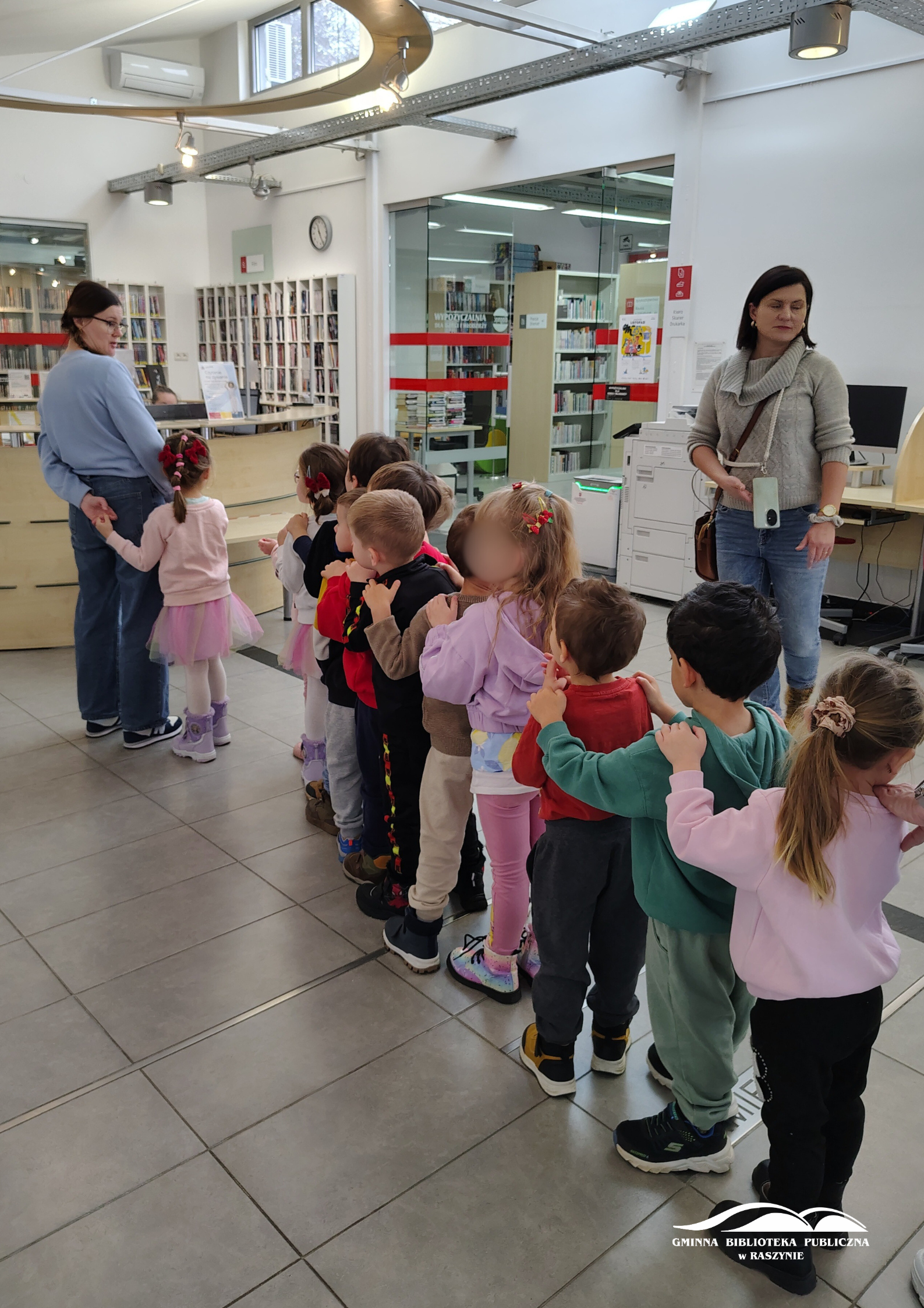 Dzieci stoją w rzędzie w holu biblioteki, witane przez bibliotekarkę