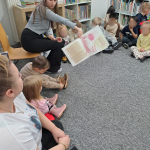 Bibliotekarka pokazuje ilustrację z kisążeczki dzieciom, które siedzą wokoło w dziale dla dzieci