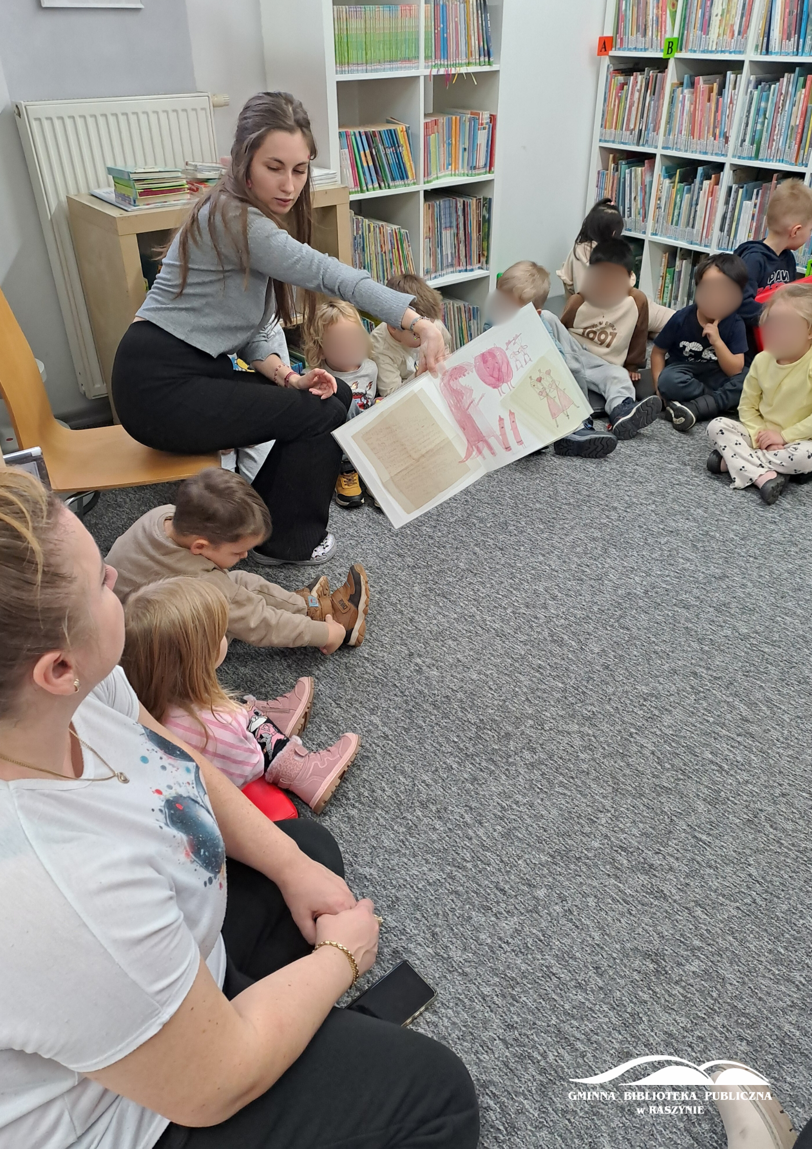 Bibliotekarka pokazuje ilustrację z kisążeczki dzieciom, które siedzą wokoło w dziale dla dzieci