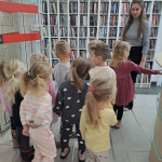 Grupa dzieci stoi przed działem z multimediami, bibliotekarka pokazuje jeden z regałów