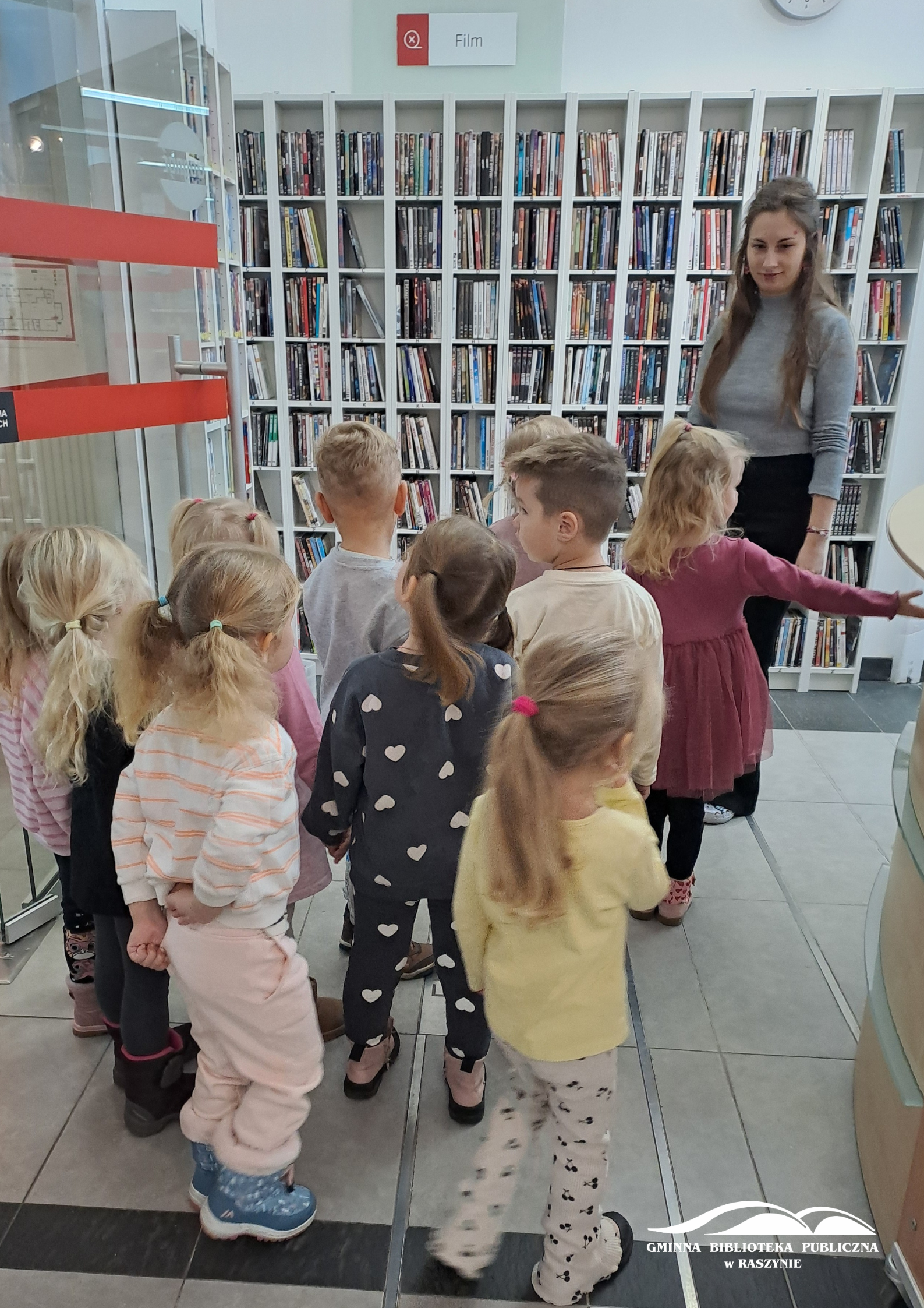 Grupa dzieci stoi przed działem z multimediami, bibliotekarka pokazuje jeden z regałów