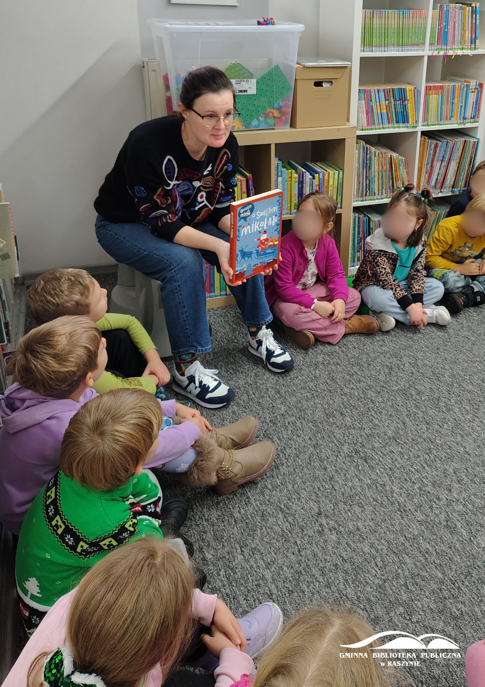 Dzieci siedzą w koło w dziale dla najmłodszych, bibliotekarka pokazuje książkę o Mikołaju