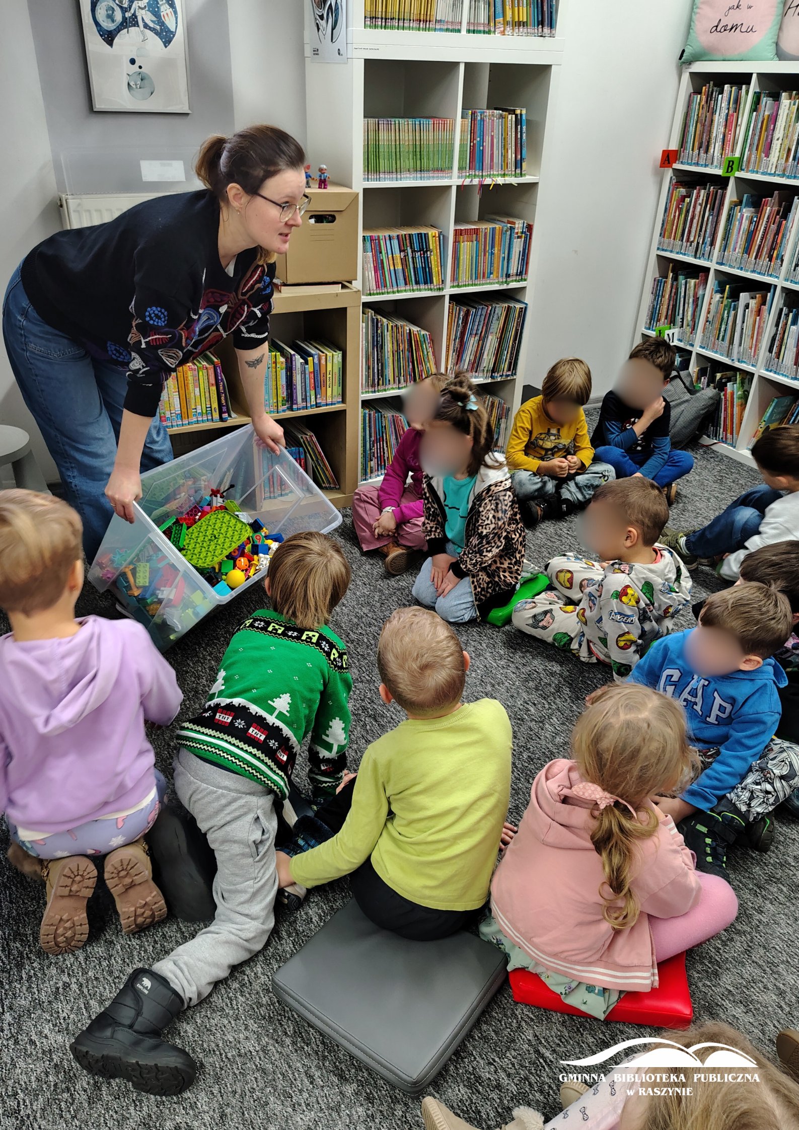 Bibliotekarka wysypuje klocki Lego z dużego pudła, dzieci siedzą na dywanie