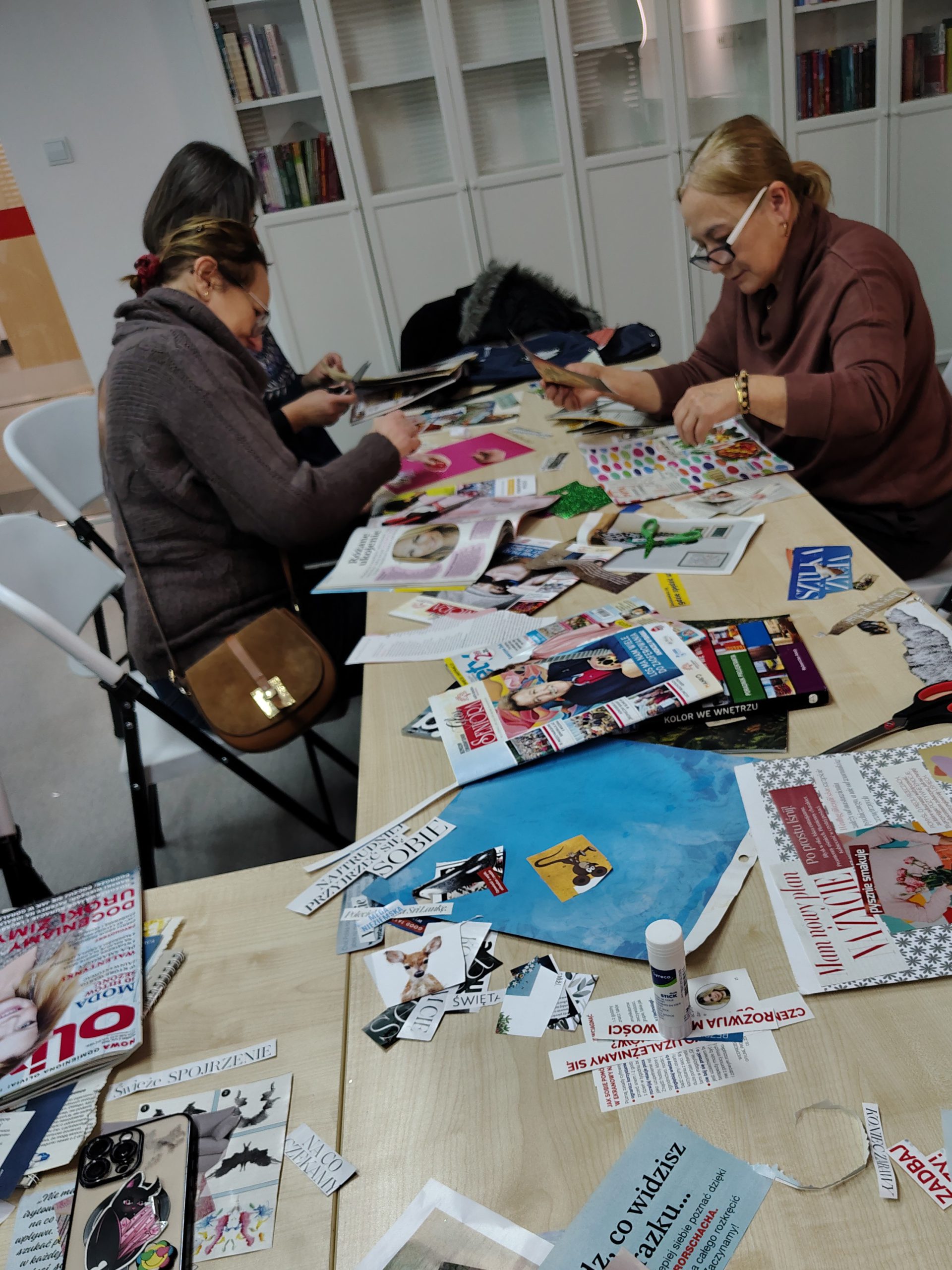 Grupa uczestników siedzi przy stołach i wycina elementy kolażu