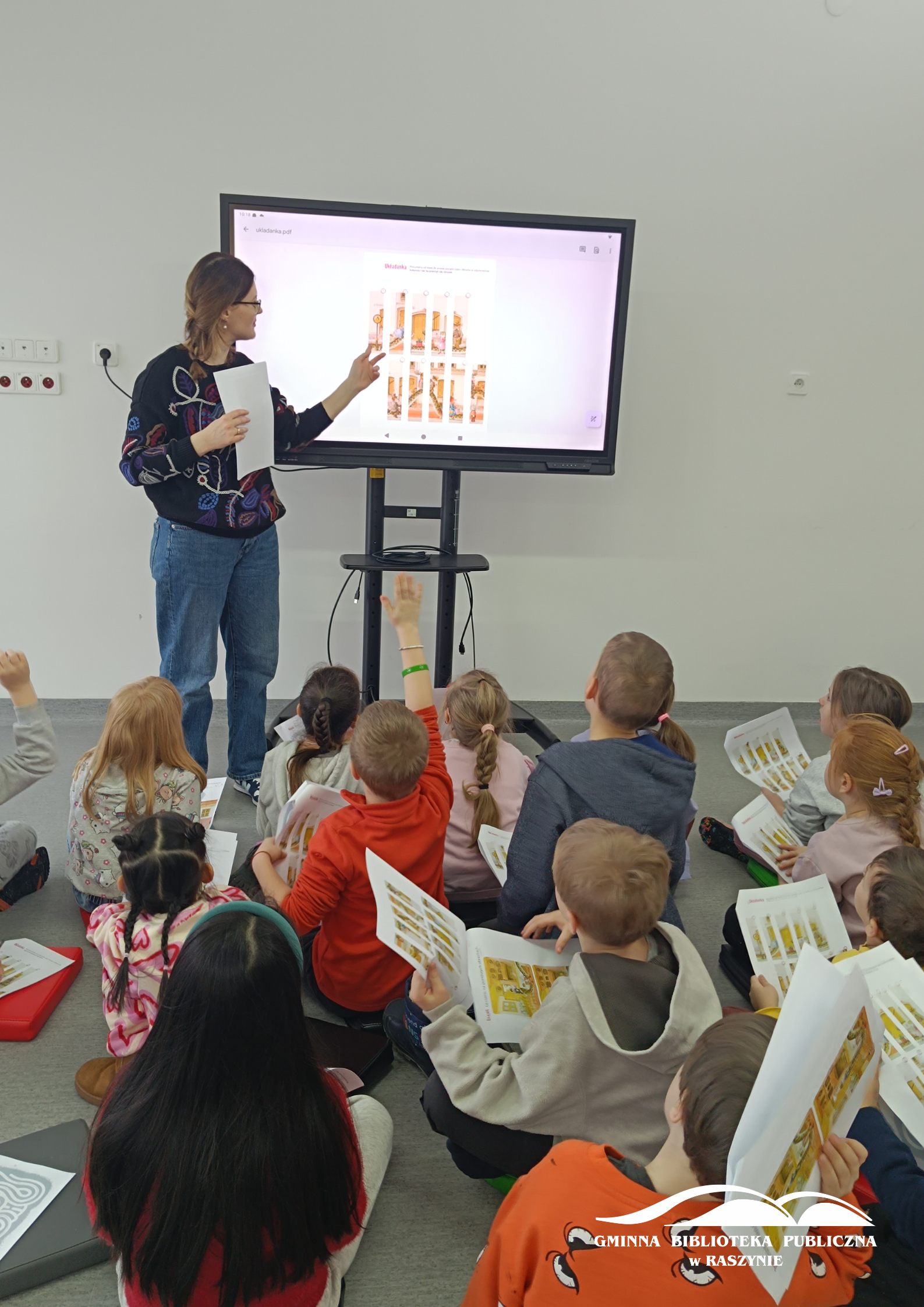 Dzieci patrzą na ekran i wspólnie z bibliotekarką rozwiązują zadanie