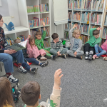 Dzieci siedzą w kole w dziale dziecięcym, mają uniesione ręce