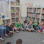 Dzieci siedzą w kole w dziale dziecięcym, widać bibliotekarkę pokazującą ilustrację