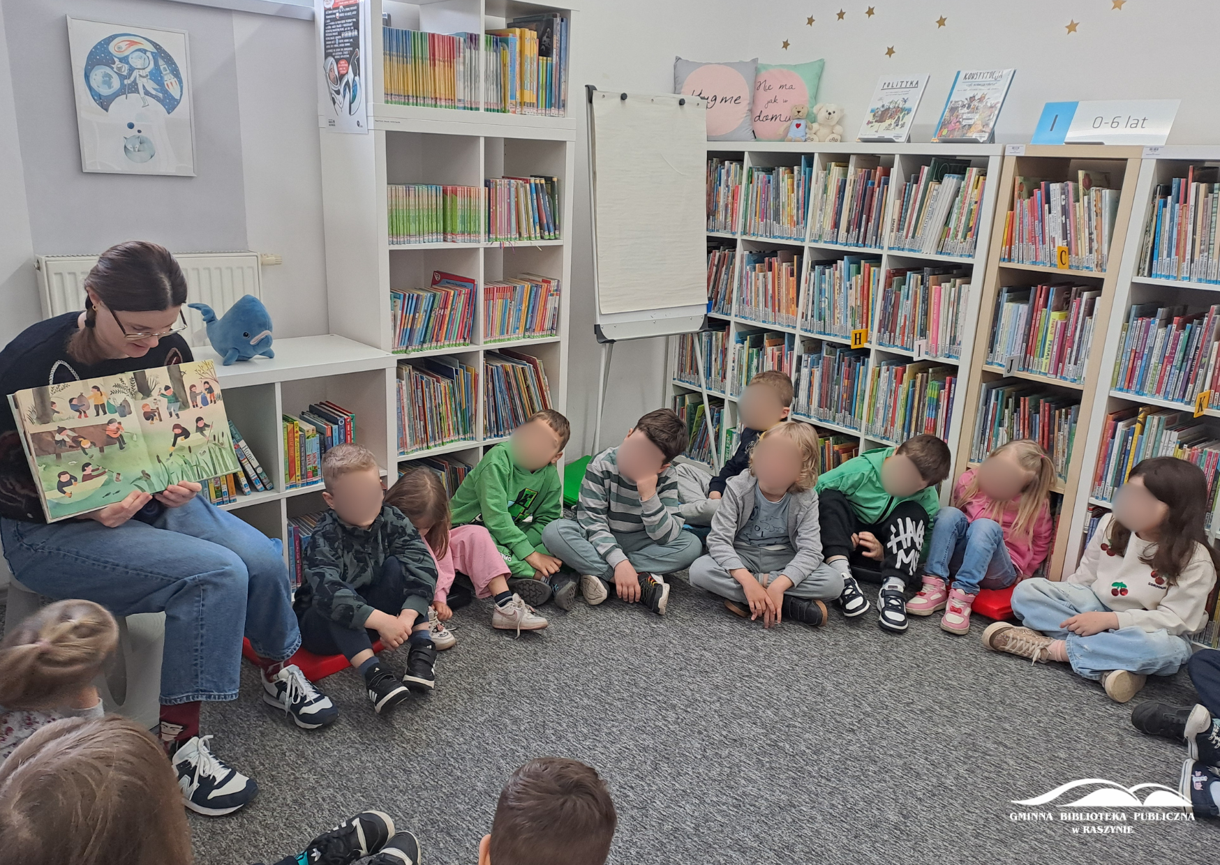 Dzieci siedzą w kole w dziale dziecięcym, widać bibliotekarkę pokazującą ilustrację