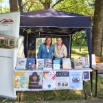 Bibliotekarki stoją na pikniku w granatowym namiocie. Widać rollup z logo biblioteki i plakaty z wydarzeniami