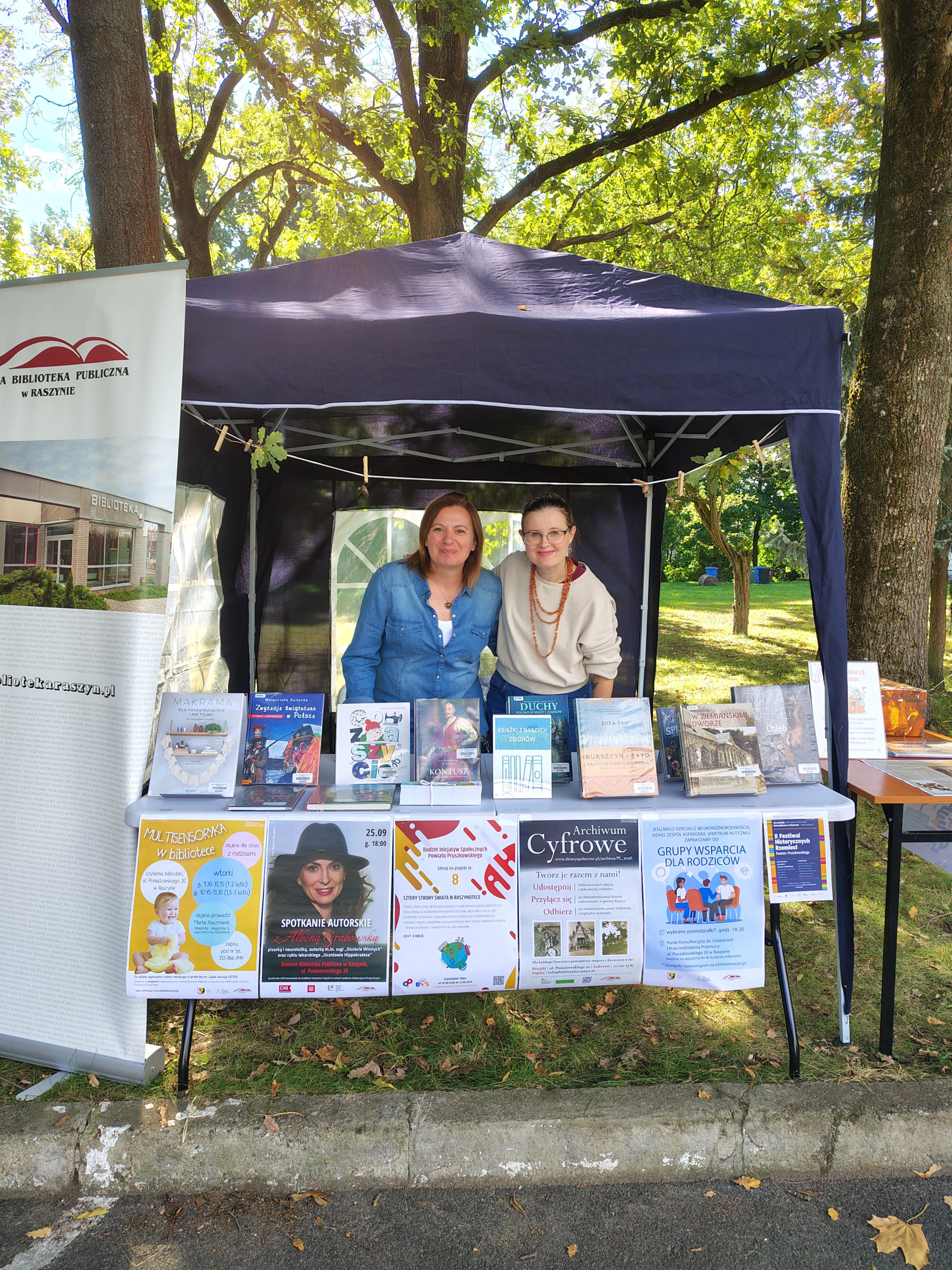 Bibliotekarki stoją na pikniku w granatowym namiocie. Widać rollup z logo biblioteki i plakaty z wydarzeniami
