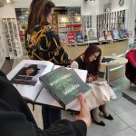 Kronika biblioteczna otwarta na stronie z plakatem reklamującycm spotkanie z Ałbeną Grabowską i wolnym miejscem na wpis autorki, na dalszym planie Autorka wpisuje dedykację w książce dla stojącej obok czytelniczki