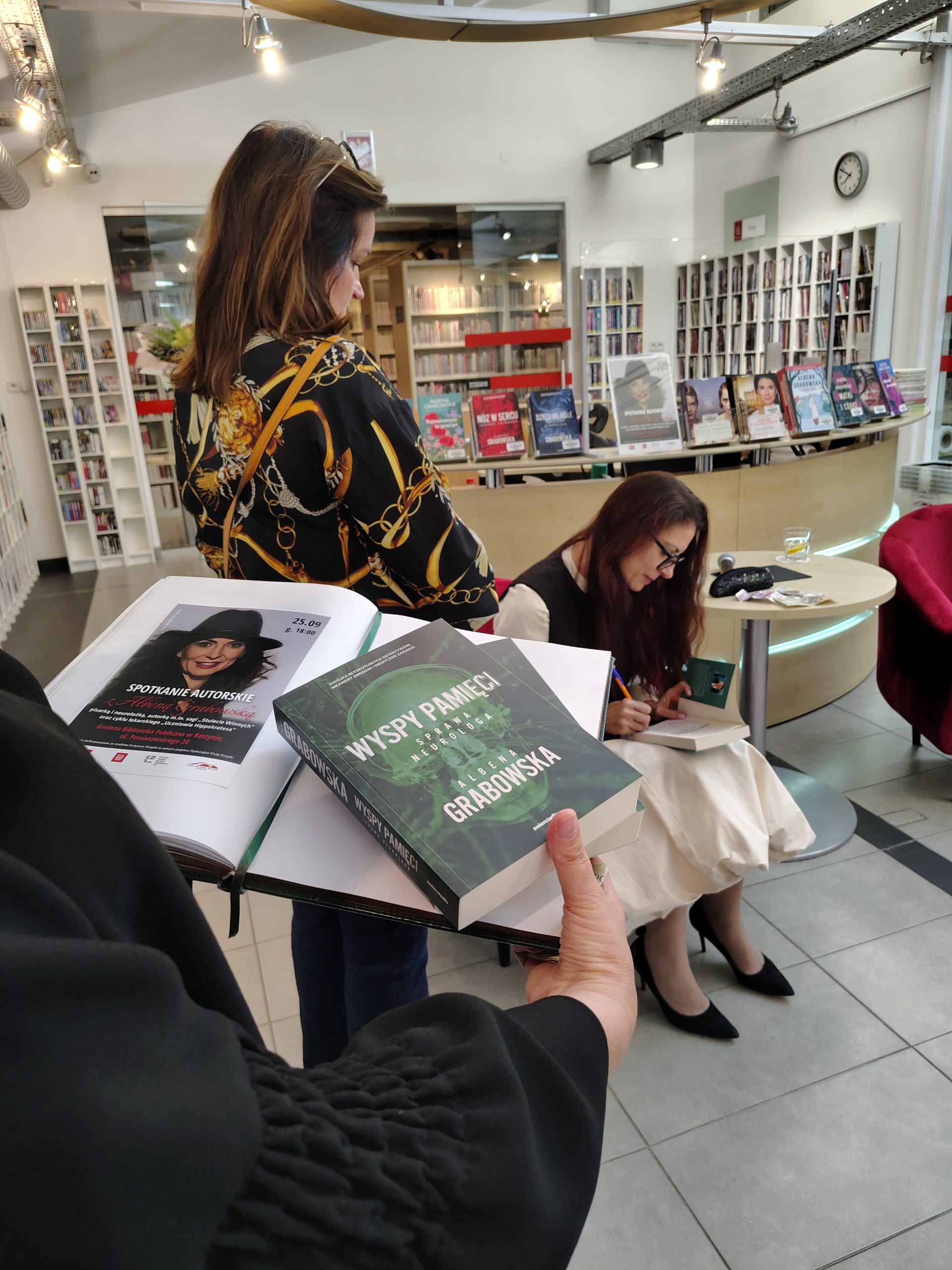Kronika biblioteczna otwarta na stronie z plakatem reklamującycm spotkanie z Ałbeną Grabowską i wolnym miejscem na wpis autorki, na dalszym planie Autorka wpisuje dedykację w książce dla stojącej obok czytelniczki